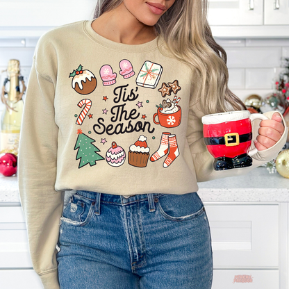 A woman wearing a sand crewneck sweatshirt with a colorful Christmas graphic that reads 'It's The Season' surrounded by holiday illustrations. She is holding a Santa-themed ceramic mug in a bright kitchen setting.