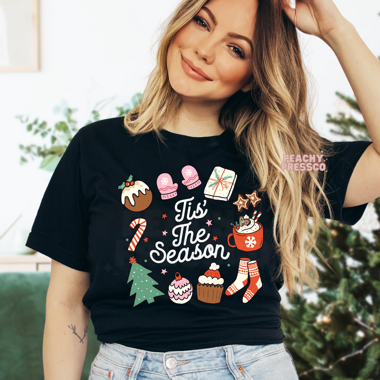 A woman wearing a black graphic t-shirt with a colorful Christmas-themed print that reads 'Tis The Season' surrounded by festive illustrations like a mug, cupcake, candy cane, and stocking.