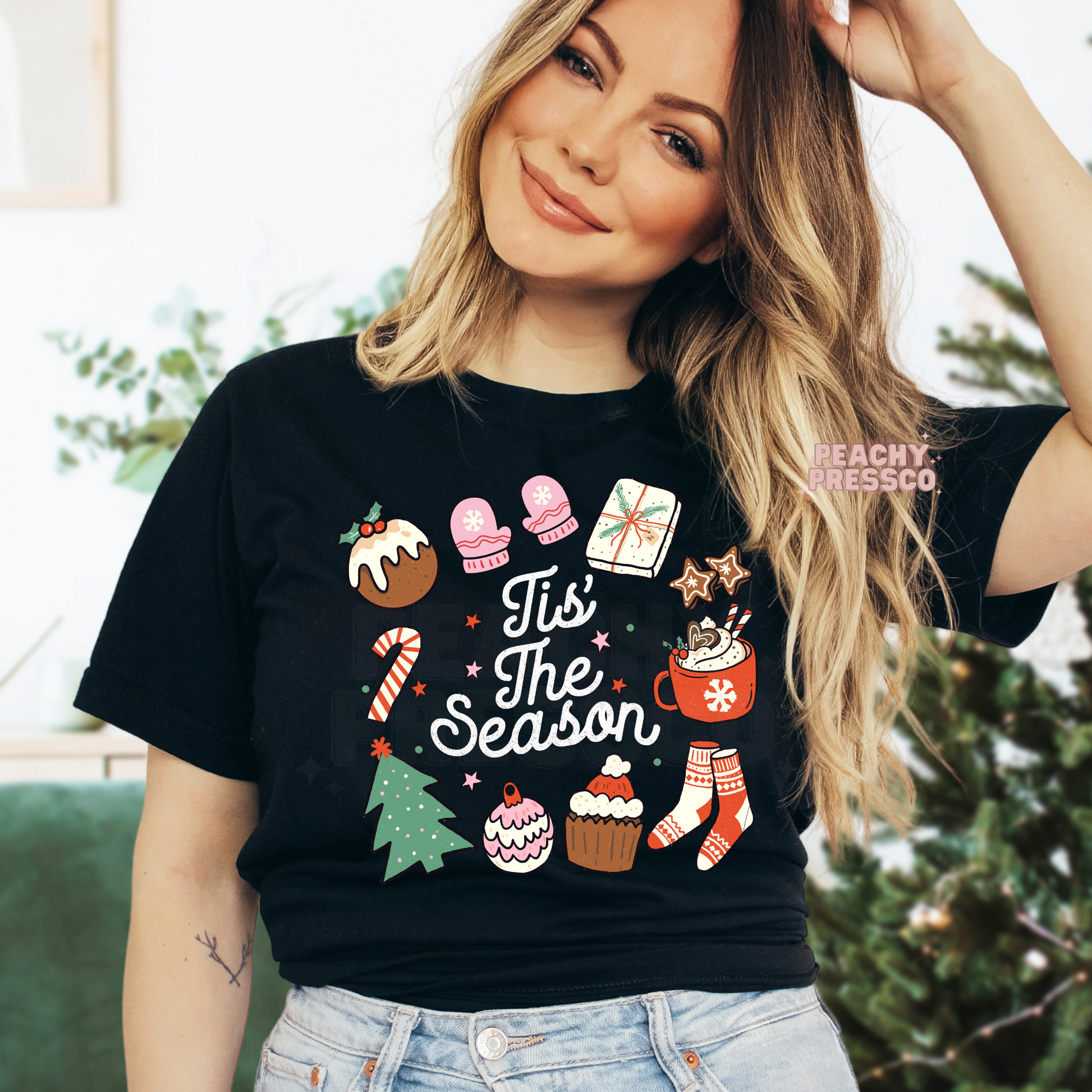 A woman wearing a black graphic t-shirt with a colorful Christmas-themed print that reads 'Tis The Season' surrounded by festive illustrations like a mug, cupcake, candy cane, and stocking.