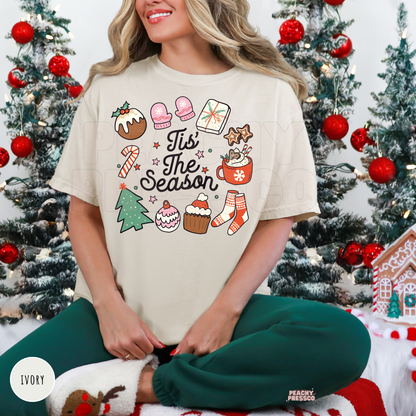 A woman wearing a ivory colored t-shirt with a colorful Christmas graphic that reads 'It's The Season' surrounded by festive illustrations, sitting cross-legged in front of decorated Christmas trees.