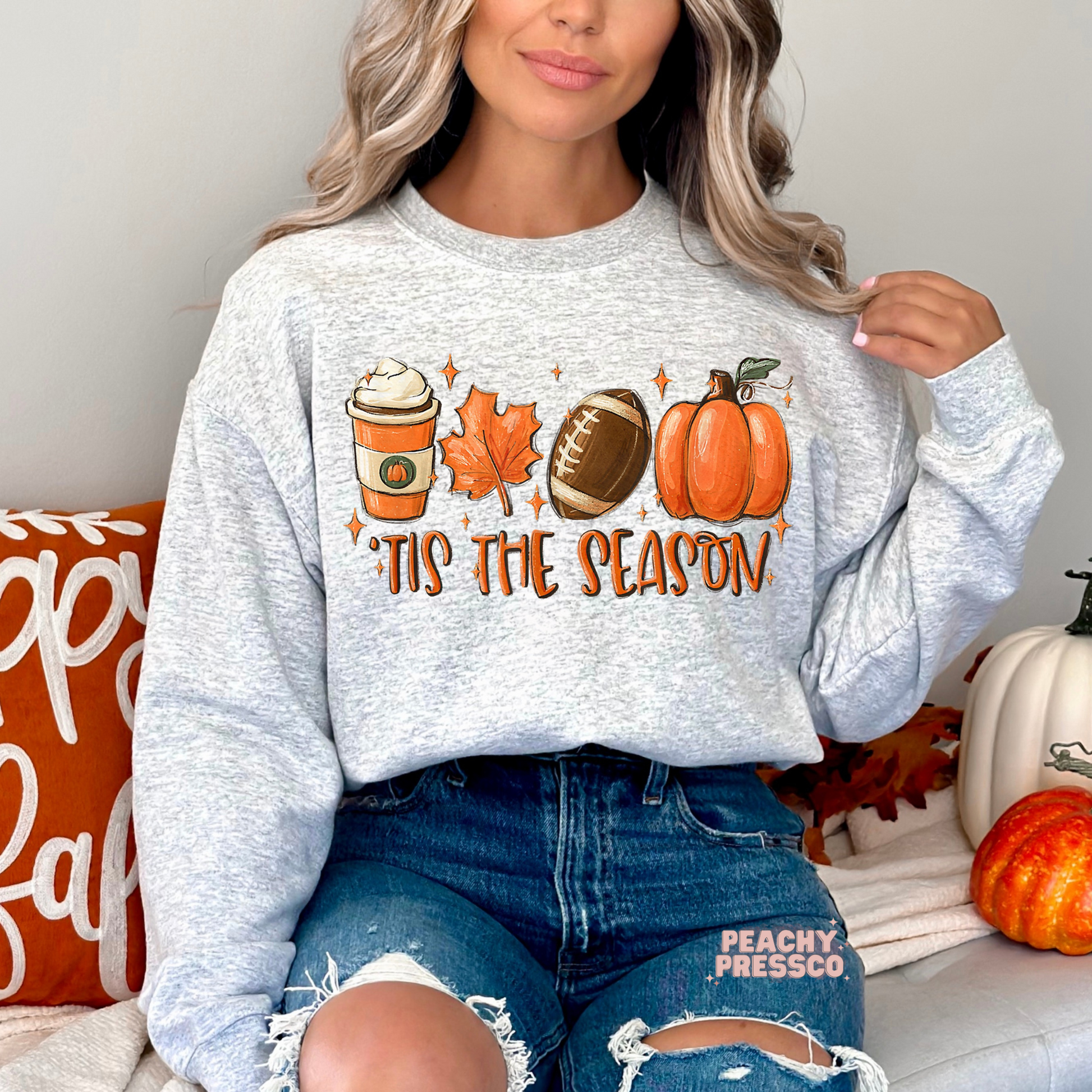 Woman wearing an ash grey crewneck sweatshirt featuring a fall football design with pumpkin spice coffee, maple leaf, pumpkin, and "'Tis The Season" text.