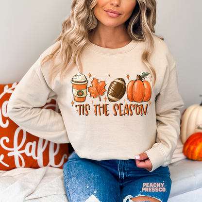 Woman wearing a sand crewneck sweatshirt featuring a "'Tis The Season" graphic with a football, pumpkin spice latte, autumn leaf, and pumpkin for fall.