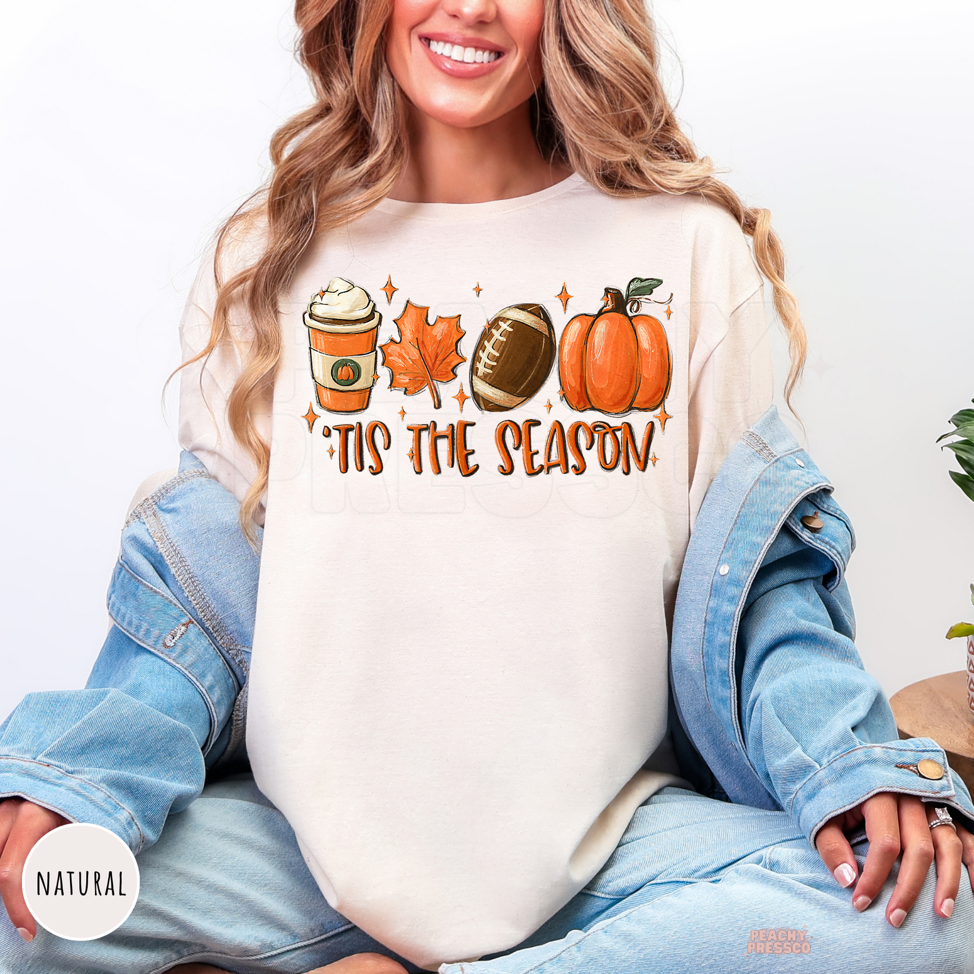 Woman wearing a natural graphic t-shirt with a cozy fall design featuring pumpkin spice coffee, football, pumpkin, maple leaf, and "'Tis The Season" artwork.