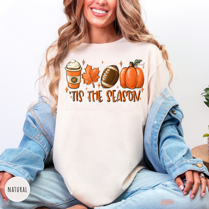 Woman wearing a natural graphic t-shirt with a cozy fall design featuring pumpkin spice coffee, football, pumpkin, maple leaf, and "'Tis The Season" artwork.