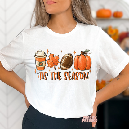 Woman wearing a white graphic t-shirt featuring a pumpkin spice coffee, football, maple leaf, pumpkin, and "'Tis The Season" fall design.
