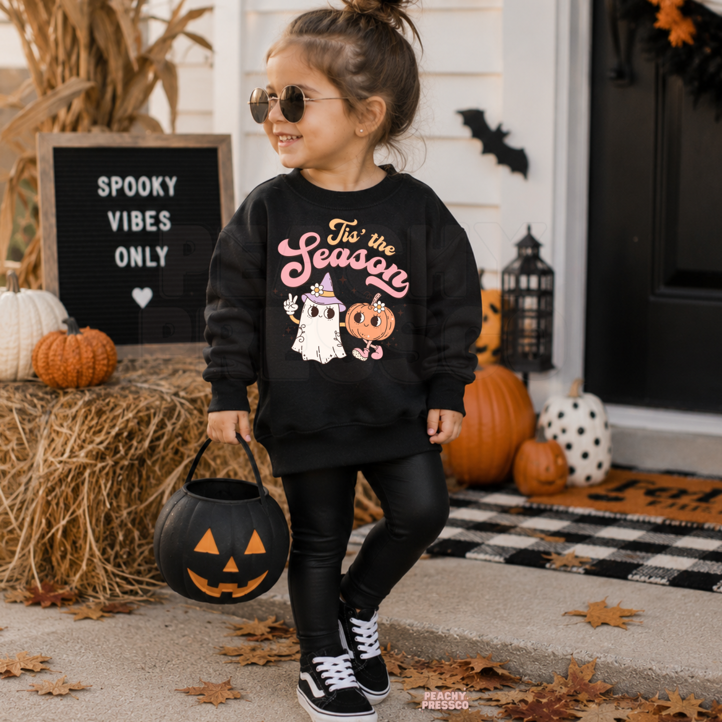 Little girl wearing a black Tis the Season Halloween sweatshirt with cute ghost and pumpkin characters while holding a pumpkin trick-or-treat bucket.
