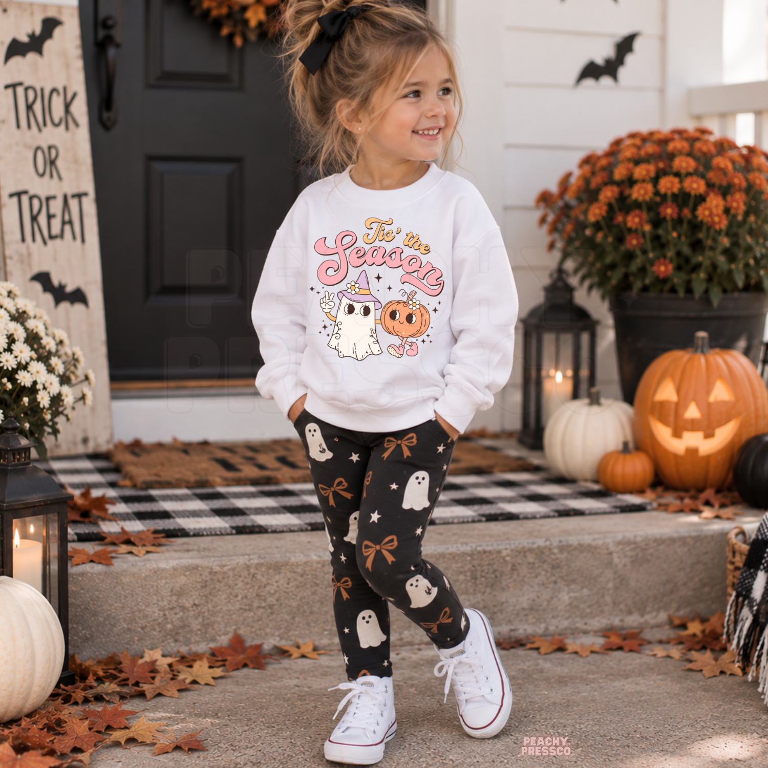 Young girl wearing a white Tis the Season Halloween sweatshirt with a girly ghost and pumpkin graphic surrounded by Halloween porch decor.