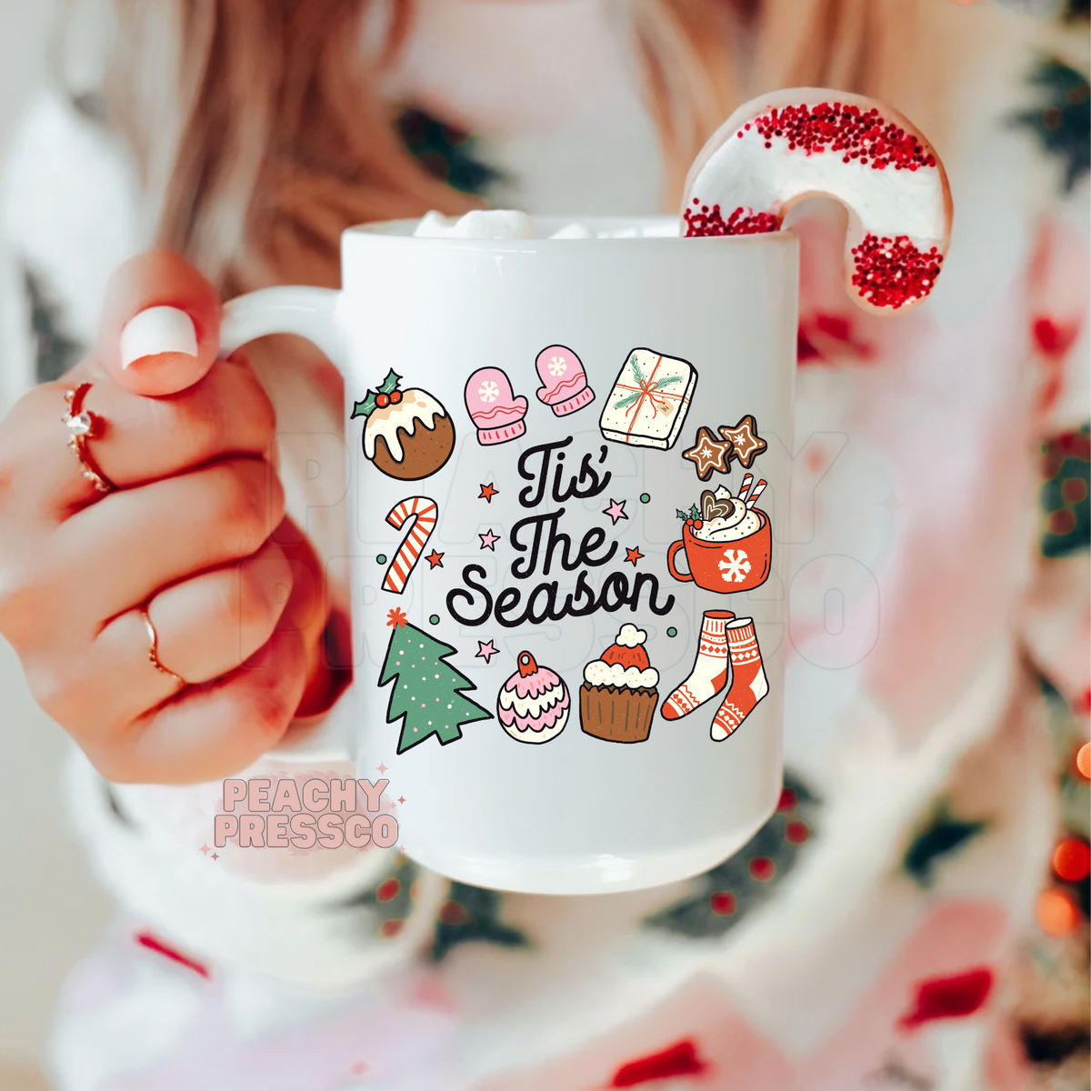 White ceramic mug printed with Tis The Season lettering surrounded by cute Christmas doodles like mittens, Christmas tree, and hot cocoa.