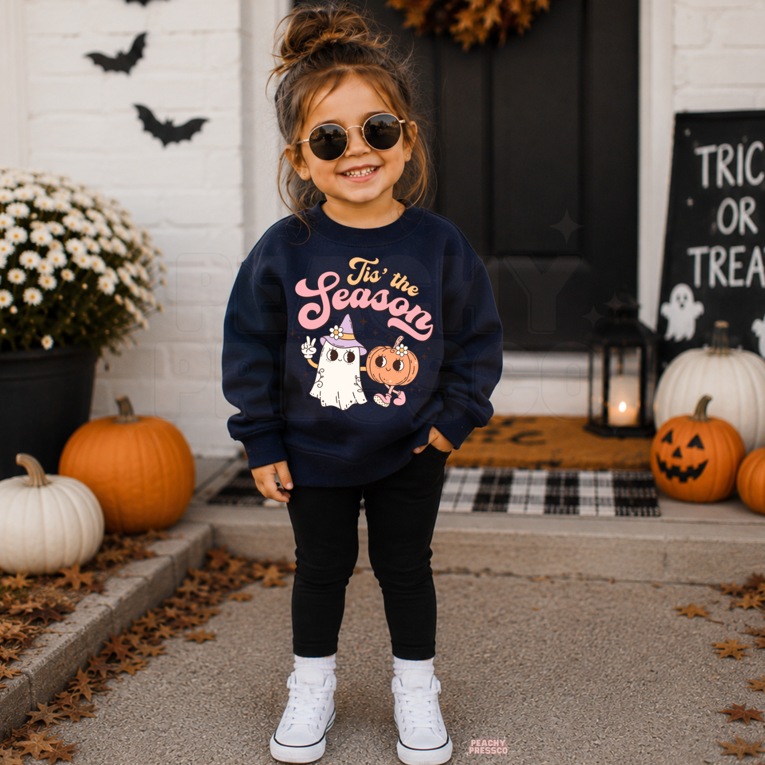 Toddler girl wearing a navy Tis the Season Halloween crewneck featuring a cute pink ghost and pumpkin design on a decorated fall porch.