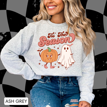 Woman wearing an ash grey crewneck sweatshirt with a retro "Tis The Season" pumpkin and ghost Halloween design.