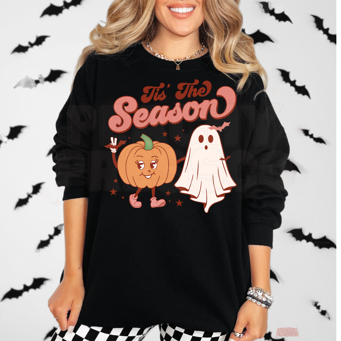Woman wearing a black crewneck sweatshirt with a retro "Tis The Season" pumpkin and ghost Halloween graphic.