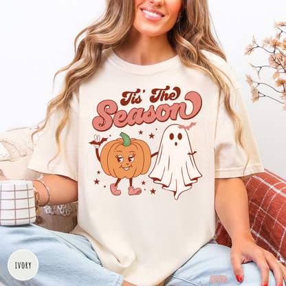 Woman wearing an ivory t-shirt with a retro "Tis The Season" pumpkin and ghost Halloween graphic design.