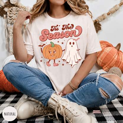 Woman wearing a natural t-shirt featuring a cute pumpkin and ghost "Tis The Season" Halloween graphic.