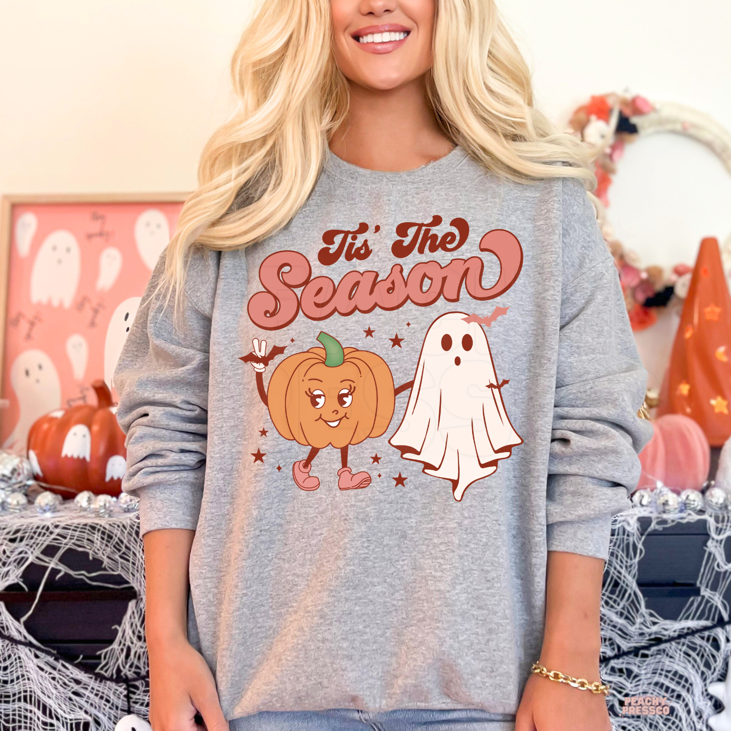 Woman wearing a sport grey crewneck sweatshirt featuring a cute retro pumpkin and ghost Halloween graphic.