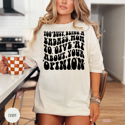 Ivory t-shirt featuring the bold "Too Busy Being a Badass Mom to Give AF About Your Opinion" graphic. Funny mom tee for everyday wear, Mother's Day, and busy moms.