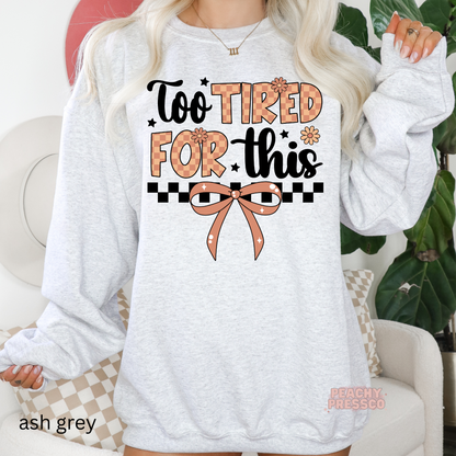 Woman wearing an ash grey sweatshirt with a funny "Too Tired For This" graphic, featuring a coquette bow, checkerboard stripe, and floral details.