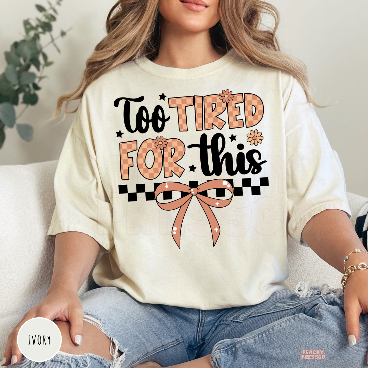 Woman wearing an ivory graphic t-shirt with a "Too Tired For This" design featuring a vintage checkerboard pattern, bow, and floral accents.