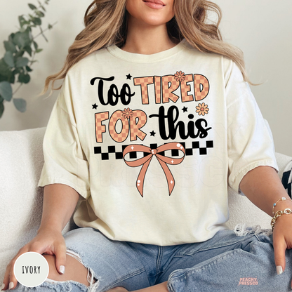 Woman wearing an ivory graphic t-shirt with a "Too Tired For This" design featuring a vintage checkerboard pattern, bow, and floral accents.