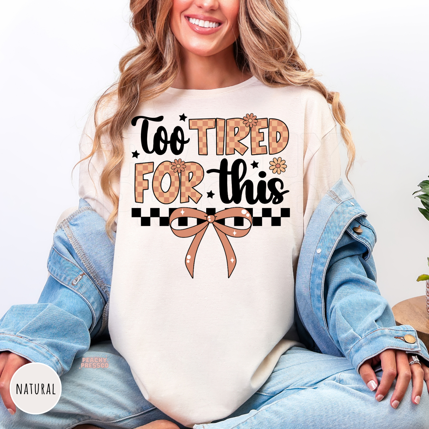 Woman wearing a natural shirt featuring a "Too Tired For This" graphic with a coquette bow, checkerboard accents, and daisy flowers.