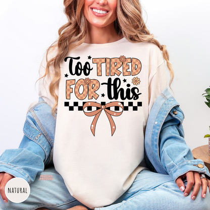 Woman wearing a natural shirt featuring a "Too Tired For This" graphic with a coquette bow, checkerboard accents, and daisy flowers.