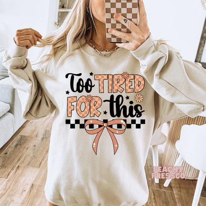 Mirror selfie of a woman wearing a sand crewneck sweatshirt featuring the "Too Tired For This" graphic with a bow and retro checkerboard details.