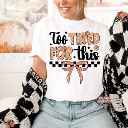 Woman wearing a white graphic t-shirt with a funny "Too Tired For This" quote, coquette bow, checkerboard design, and daisy flowers.