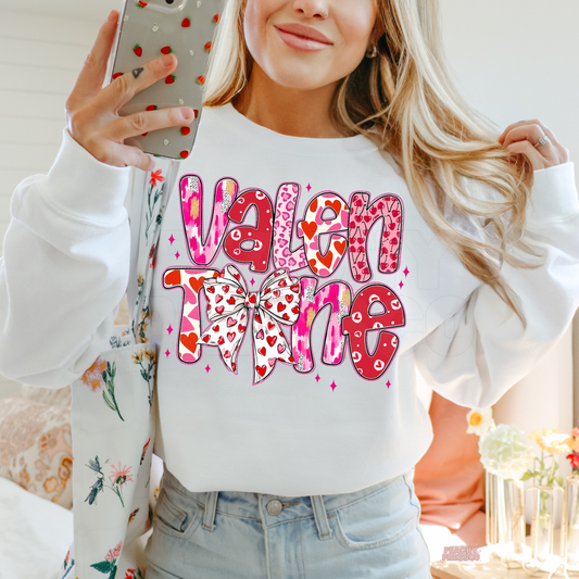 White Valentine's Day t-shirt featuring a colorful stacked "Valentine" graphic with a heart-print coquette bow.
