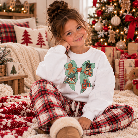 A young girl sits on a fluffy cream rug in a cozy Christmas living room, wearing a white long-sleeve pajama top with a green bow and gingerbread cookie graphic, paired with red and green plaid pajama pants.