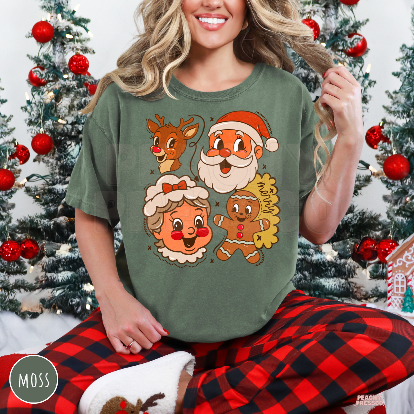 Woman wearing a moss green holiday t-shirt featuring retro Santa, Mrs. Claus, reindeer, and gingerbread man in front of Christmas trees.