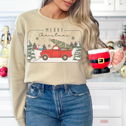 Woman in a kitchen holding a Santa boot mug wearing a sand crewneck sweatshirt with a vintage red Christmas truck graphic.