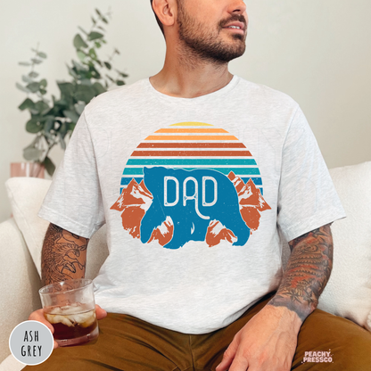 Ash Grey unisex t-shirt with a vintage sunset, mountain scenery, and blue Dad bear graphic, a rustic Father's Day shirt for dads.