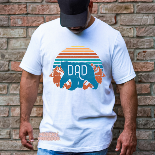White unisex t-shirt featuring a retro sunset, mountain landscape, and blue Dad bear graphic, a unique Father's Day gift for nature-loving dads.