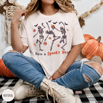Woman wearing a natural Halloween graphic tee with dancing skeletons, pumpkins, bats and "Have a Spooky Day" design.