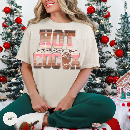 Woman sitting cross-legged wearing an ivory retro Hot Cocoa Season t-shirt with green holiday pants in front of mini Christmas trees.