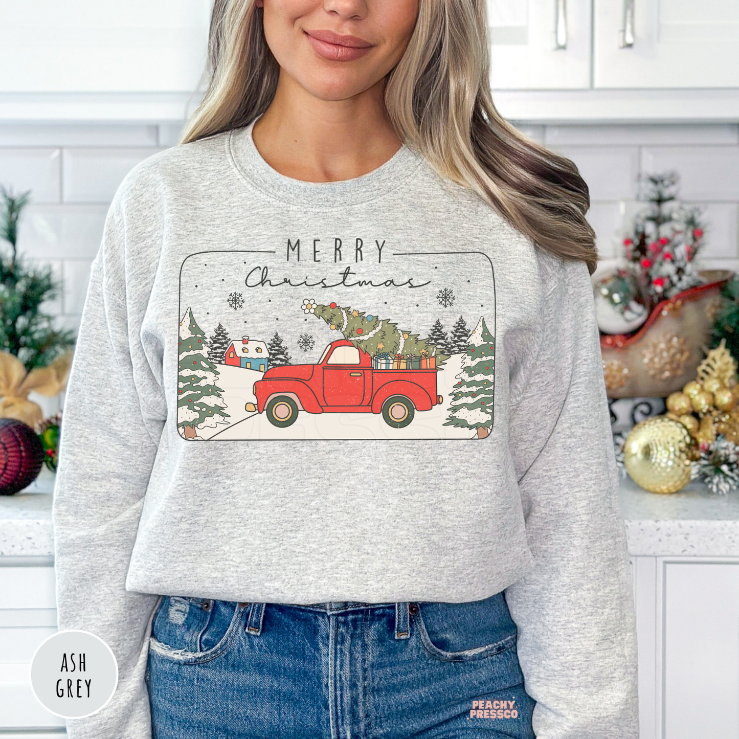Woman wearing an ash grey crewneck sweatshirt featuring a vintage red pickup truck carrying a Christmas tree with Merry Christmas text.
