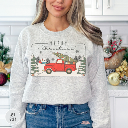 Woman wearing an ash grey crewneck sweatshirt featuring a vintage red pickup truck carrying a Christmas tree with Merry Christmas text.