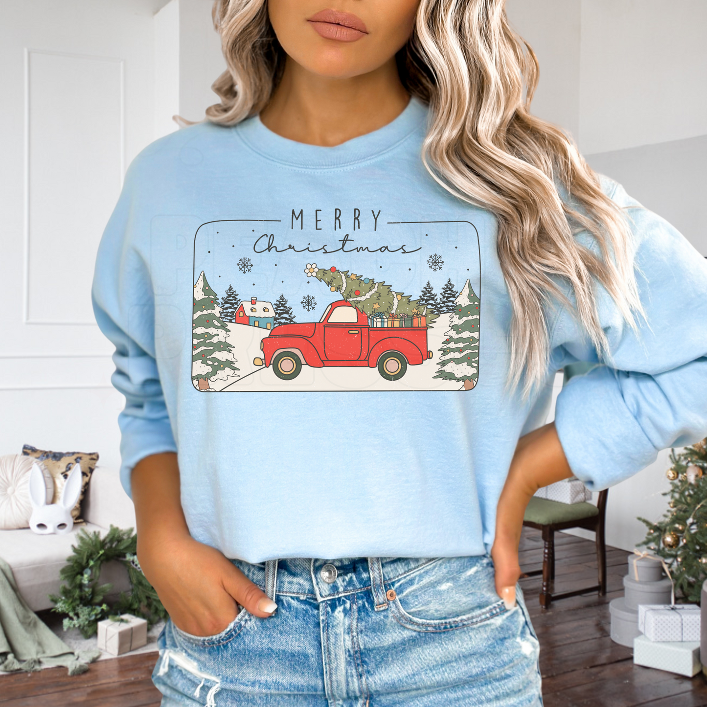 Women with hands in jeans pockets wearing a light blue crewneck sweatshirt with a Merry Christmas vintage red tree truck illustration.