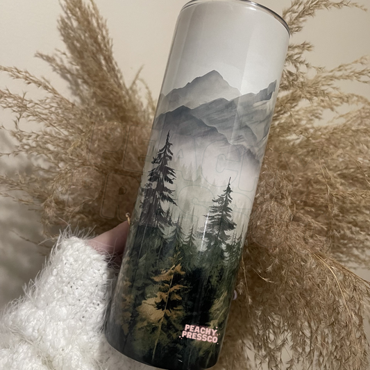 20oz skinny tumbler featuring a watercolor mountain landscape with evergreen forest and misty wilderness design.