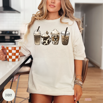 Woman wearing an ivory t-shirt featuring a western coffee lover design with four rustic coffee drink illustrations in neutral brown tones.