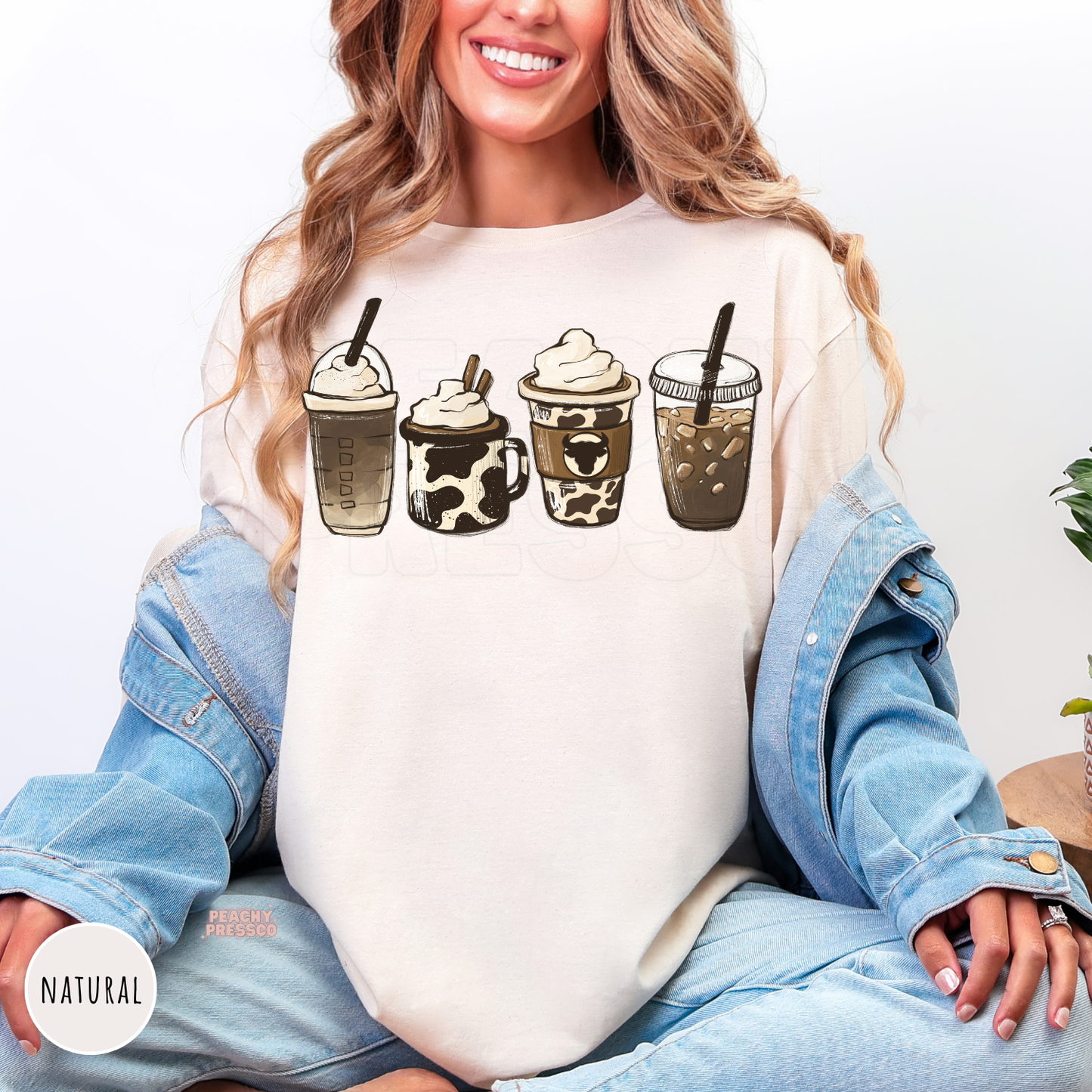 Woman wearing a natural shirt featuring four western-inspired coffee drinks with cow print accents, including iced coffee, latte, and frappé illustrations.