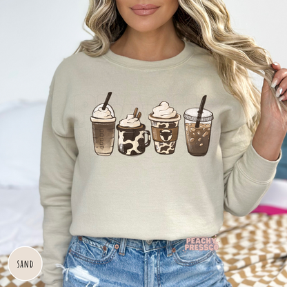 Woman wearing a sand sweatshirt with a western coffee drinks graphic showcasing cow print coffee cups and iced coffee illustrations.