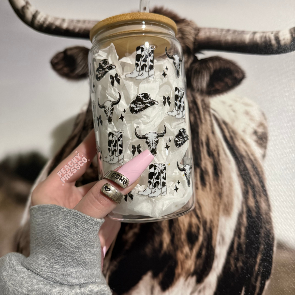 Clear glass cup featuring repeating western cowgirl boots, cow skulls, hats, bows, and stars with bamboo lid and straw.