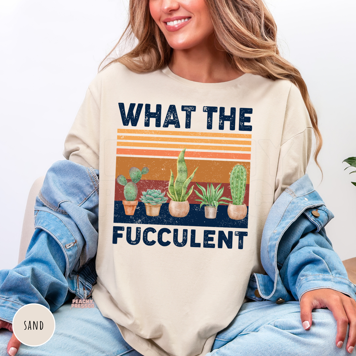 Sand crewneck sweatshirt with a humorous "What the Fucculent" design featuring potted succulents and cacti over a distressed retro sunset. Cozy botanical sweatshirt for gardeners and plant lovers.