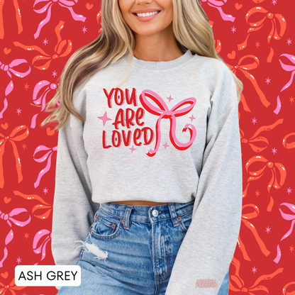 Ash grey crewneck sweatshirt displaying a cute "You Are Loved" Valentine's graphic with a pink coquette ribbon bow.