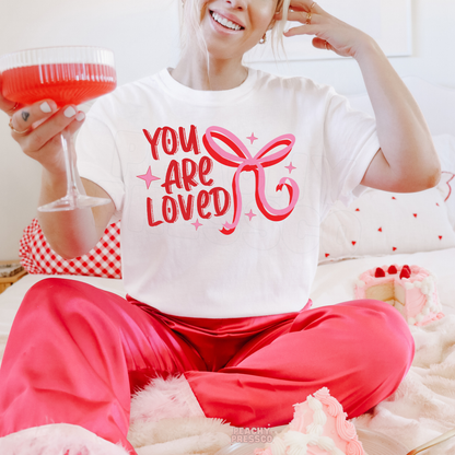 White oversized Valentine's lounge shirt featuring a retro "You Are Loved" design with a pink bow and festive accents.