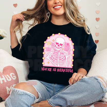 Black sweatshirt featuring a vibrant pink skeleton selfie graphic with heart eyes, vanity lights, and the quote "You're So Beautiful."