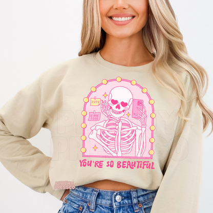Sand sweatshirt displaying a cute gothic pink skeleton taking a mirror selfie with heart eyes and "You're So Beautiful" Valentine's graphic.
