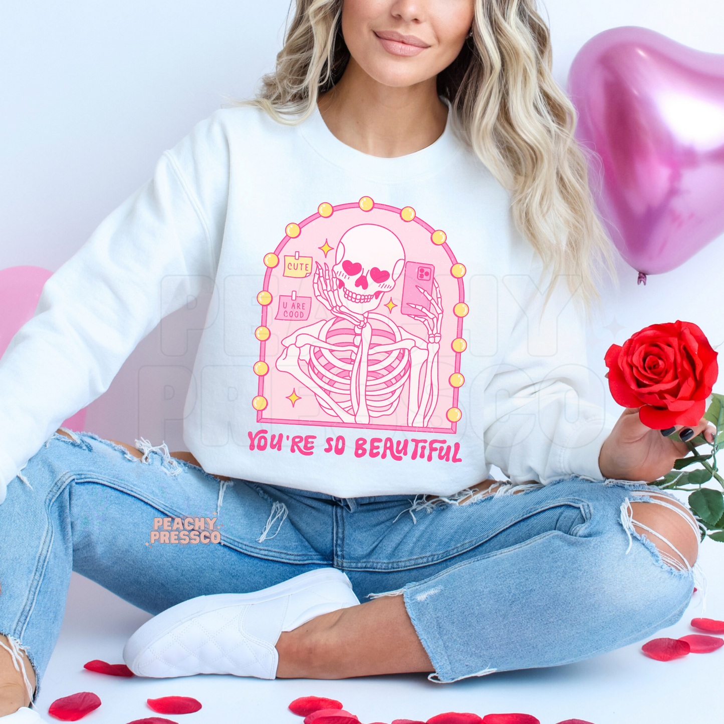 White sweatshirt displaying a pink skeleton taking a mirror selfie with heart-shaped eyes and the phrase "You're So Beautiful" in a trendy Valentine's aesthetic.