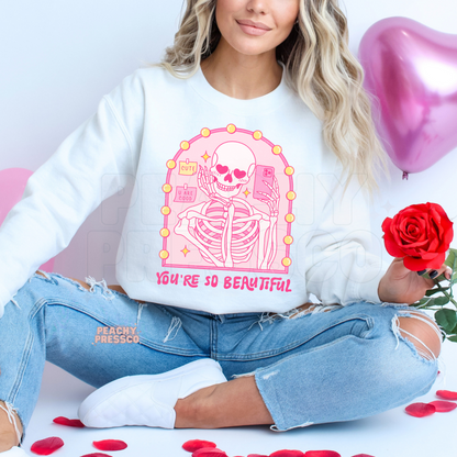 White sweatshirt displaying a pink skeleton taking a mirror selfie with heart-shaped eyes and the phrase "You're So Beautiful" in a trendy Valentine's aesthetic.